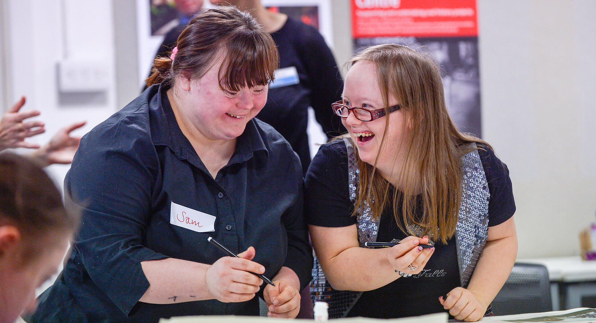 Photo of two young women with down's syndrome laughing and looking at one another. They are holding pens, and are in the middle of an activity.