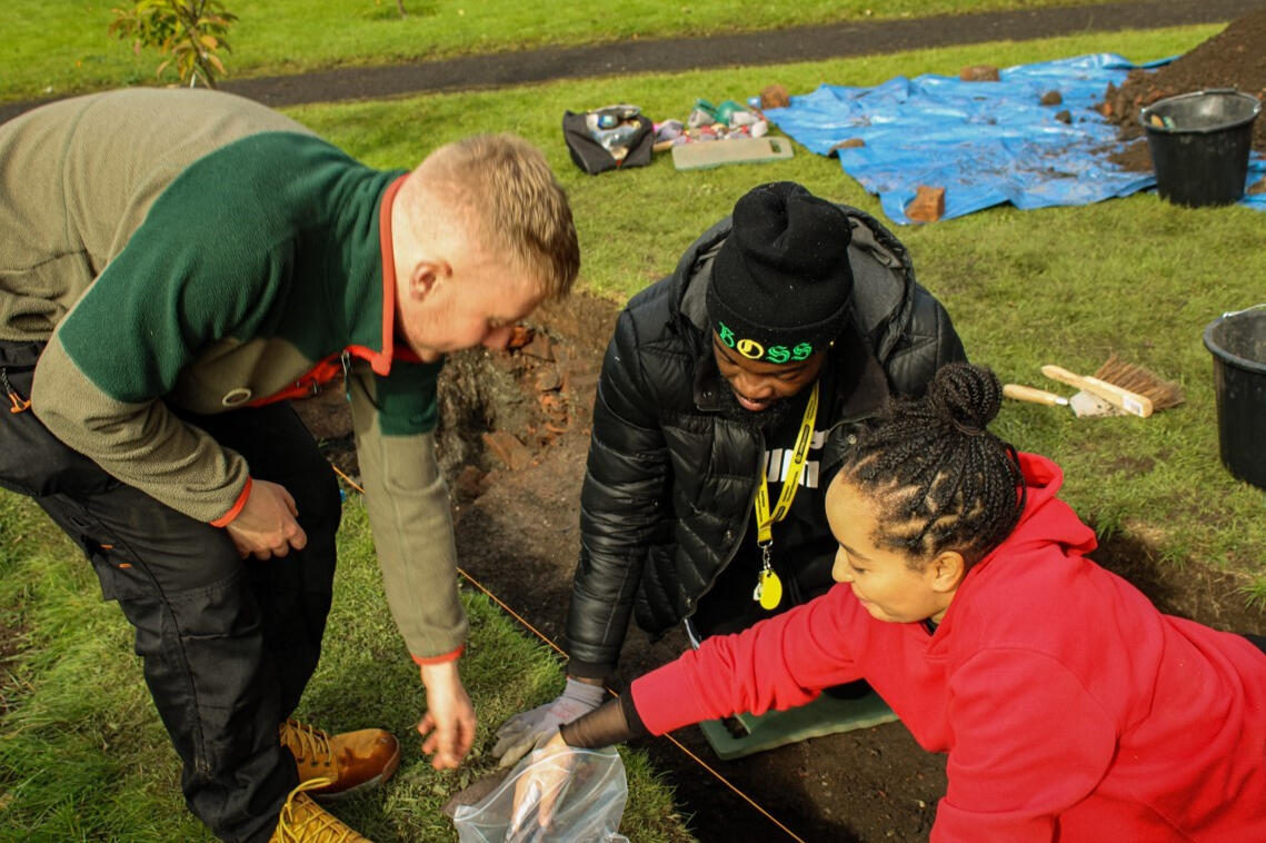 Photo of a white man, a black man, and a black woman working together in an archaeologist's trench. The black woman is placing an artefact inside of a ziplock pouch.