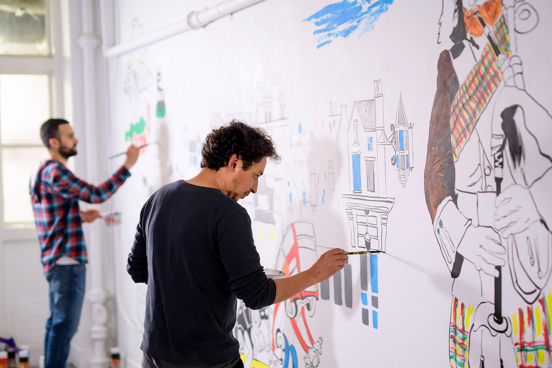 Photo of two men in the process of painting a mural on an interior wall. The mural features particularly Scottish elements, such as someone playing the bagpipes, and Scottish architecture.