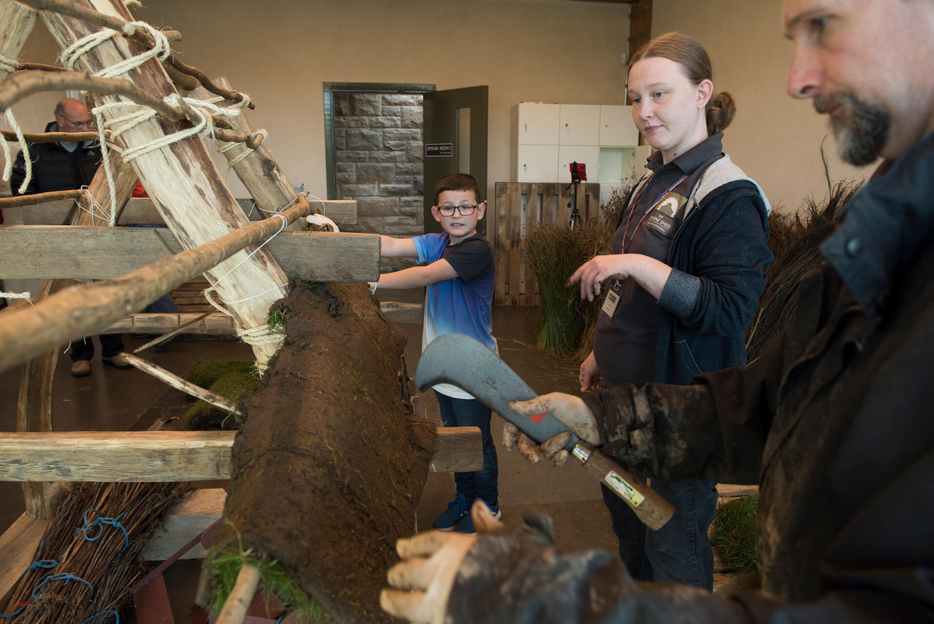 Photo of an older man demonstrating to a teenage girl and a young boy how to apply turf soil thatching to a wooden roof scaffold.