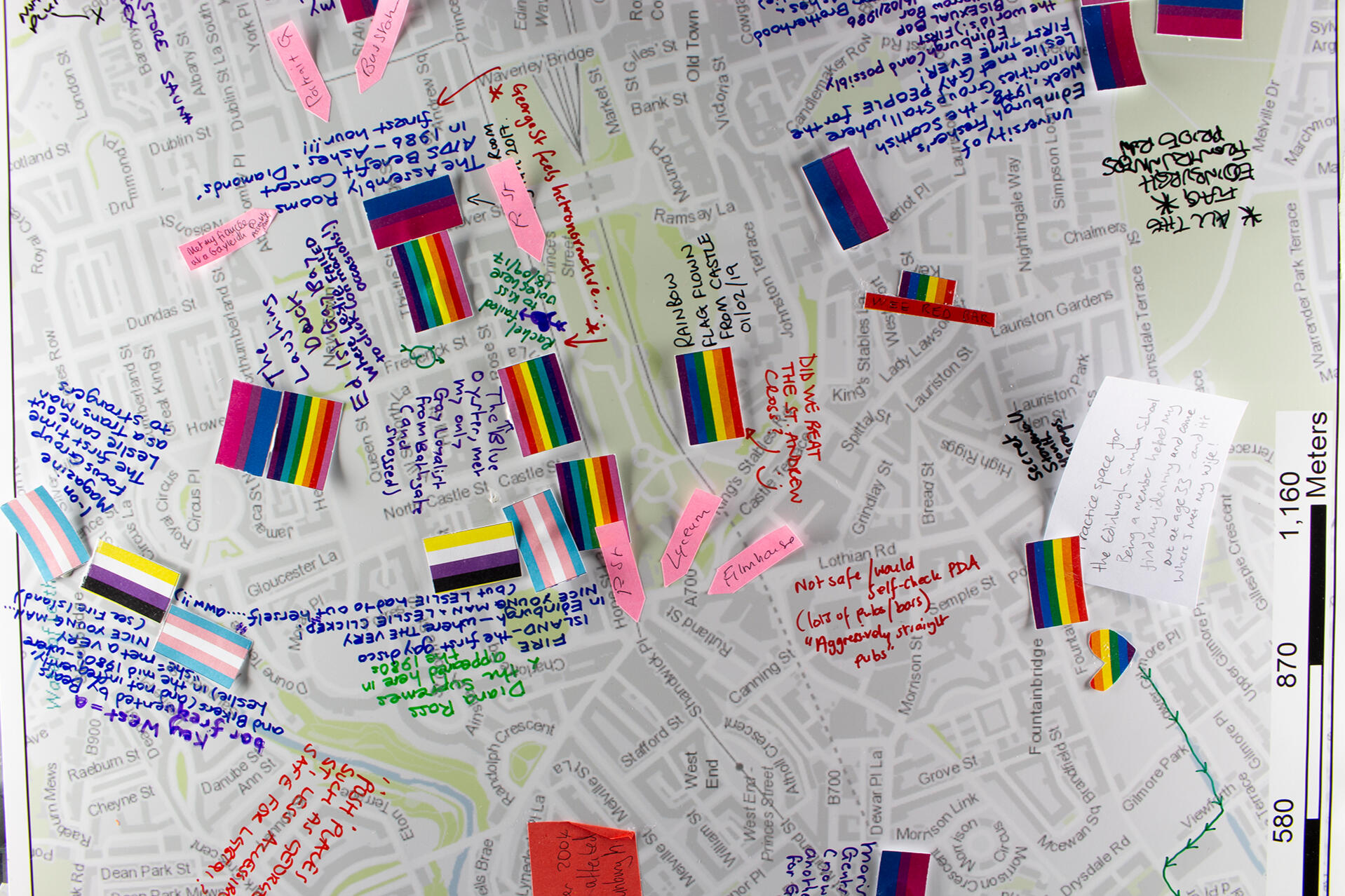 Photo of a street map of Edinburgh. LGBT, Trans, Non-binary and other queer flags have been pinned onto the map, alongside handwritten notes and sticky arrows pointing at specific locations. Comments on the map discuss queer experiences in the city.