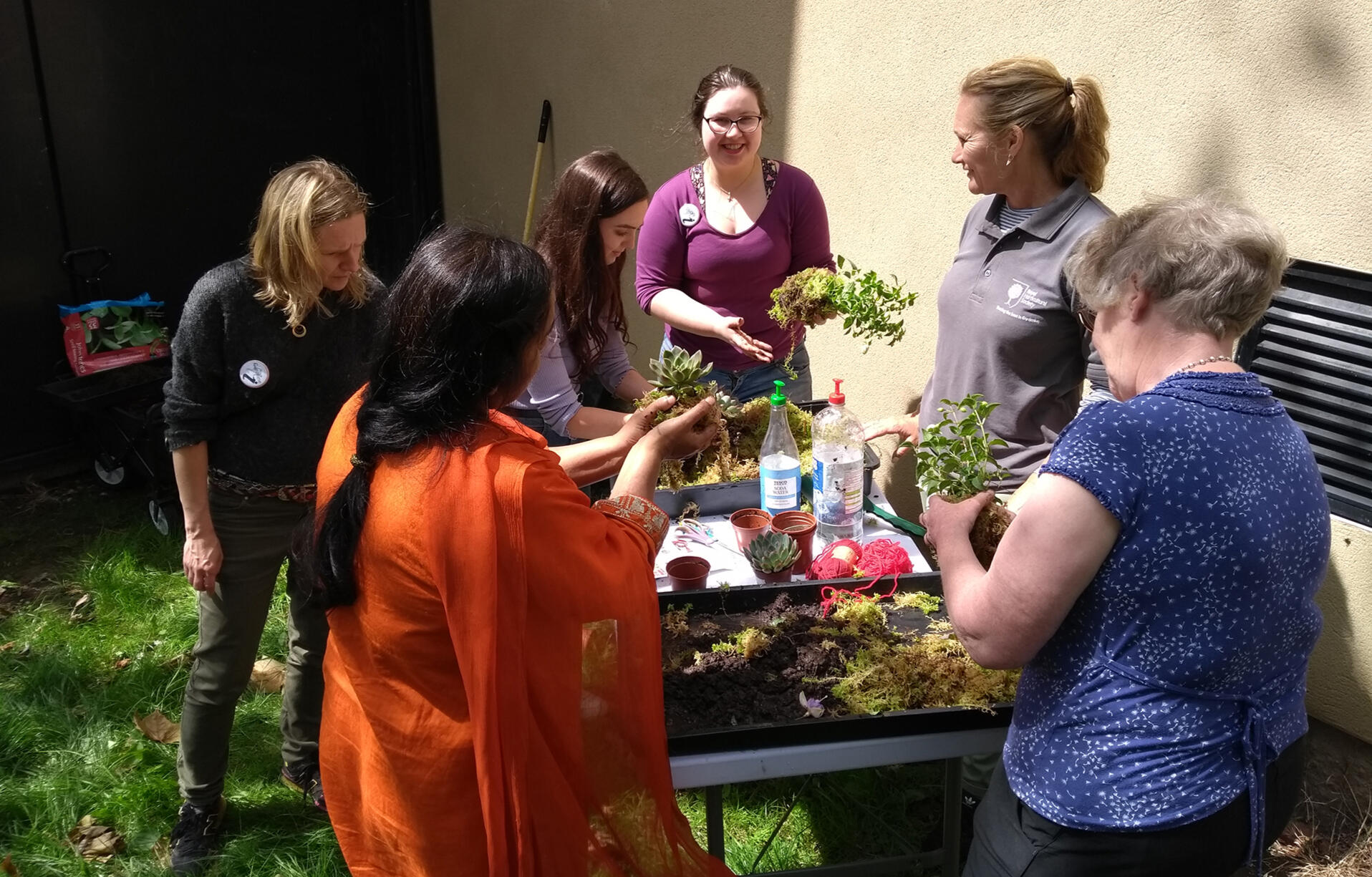 Photo of a group of women standing in a circle around a table full of plants, plant pots and gardening tools. They are chatting and laughing.